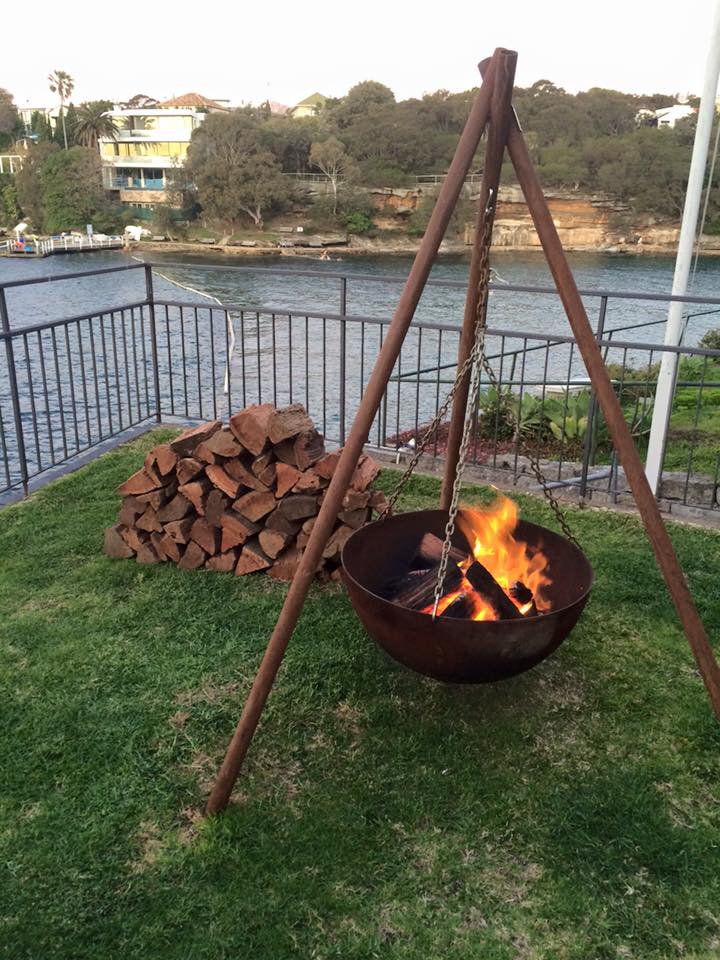 A cast iron bowl with a wood fire burning in it, hung on chains from a three-legged Tripod on a harbourside lawn beside a stack of firewood