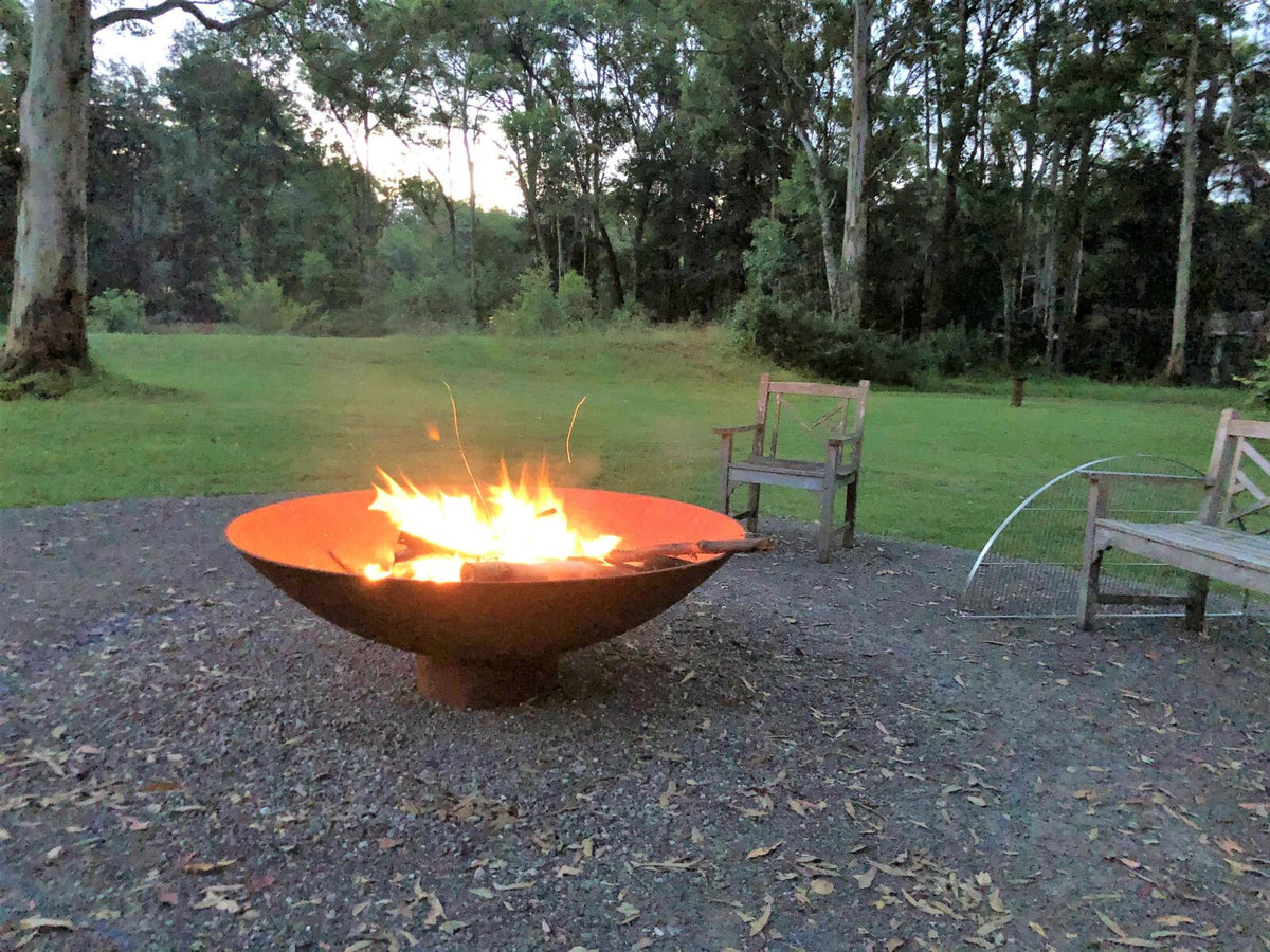 A cast iron Cauldron with a fire going on a gravel pad, with timber benches and gum trees around it