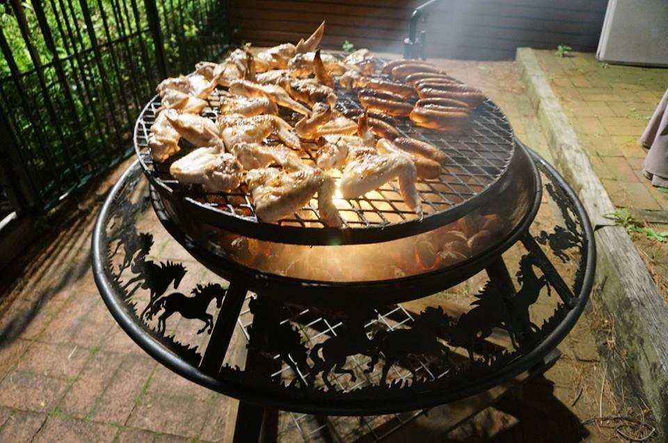 Chicken wings and sausages on the grill of a BBQ / Fire Pit at dusk, coals glowing underneath and galloping horses cut into the rim