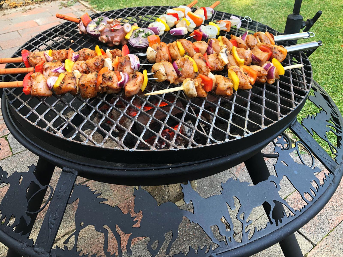 Chicken and vegetable kebabs on the grill of a BBQ / Fire Pit over glowing coals, with horses and riders cut into the rim, on a brick patio