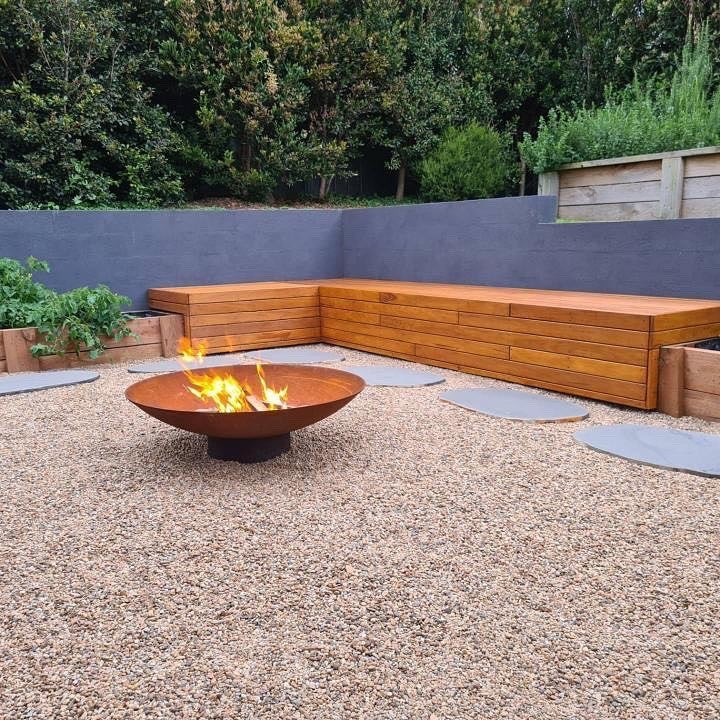 The Cauldron 800 mm with a fire going on a gravel courtyard, in front of a built-in timber bench