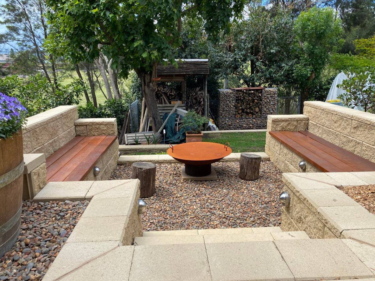 A rust-brown cast iron Cauldron in a sunken pebble courtyard, with timber bench seats, log stools and a firewood stack behind