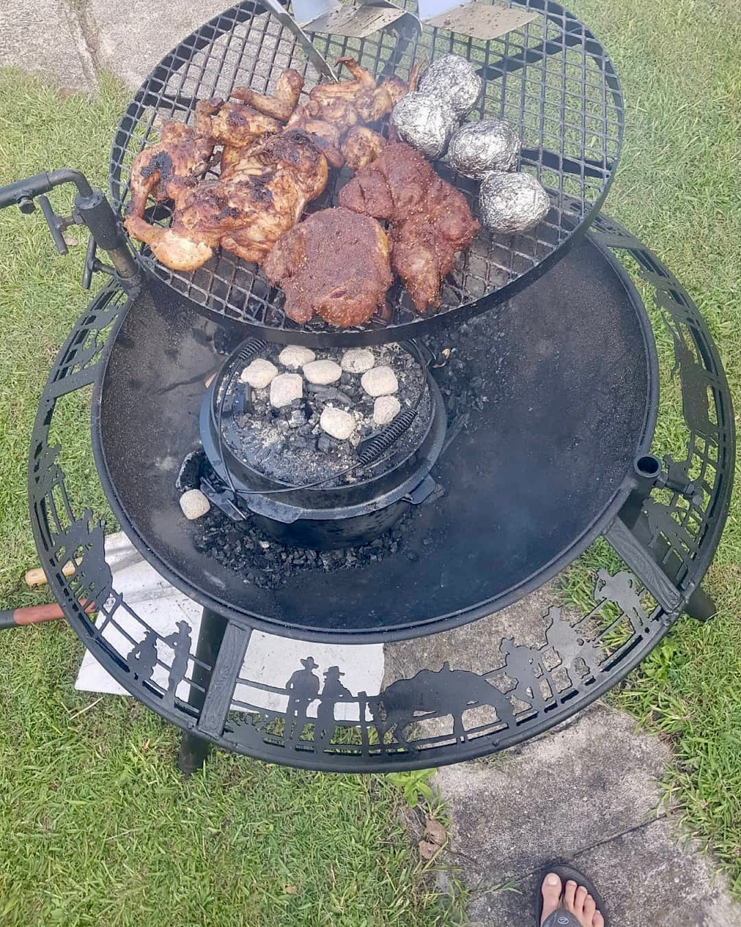 The grill of a BBQ / Fire Pit swung out to the side and loaded with chicken, steak and foil potatoes, a camp oven sitting in the coals
