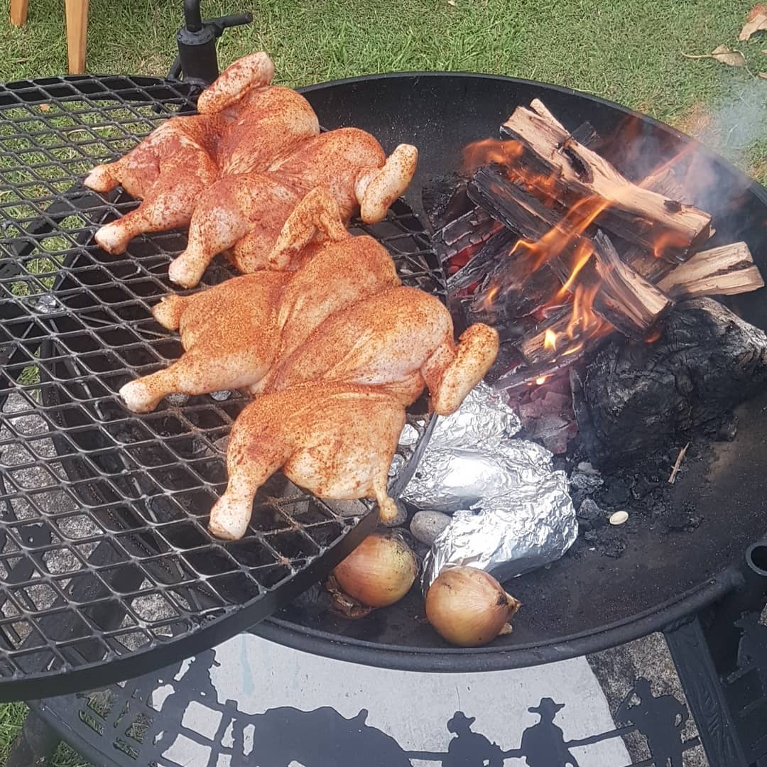 Four butterflied chickens on the grill of a BBQ / Fire Pit, a wood fire burning beside them and foil potatoes and onions in the coals