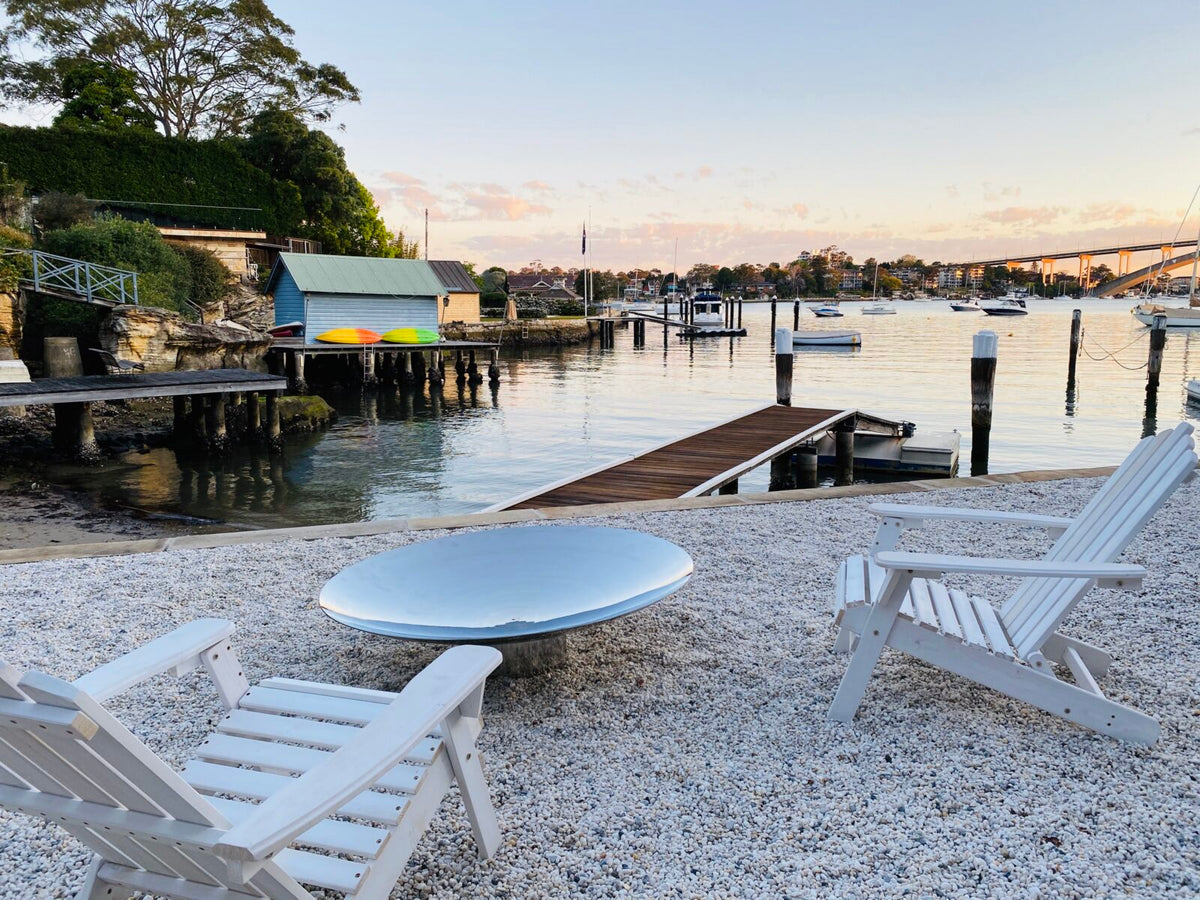 A shallow stainless steel Cauldron on white gravel by the water at dusk, with white Adirondack chairs and a jetty behind