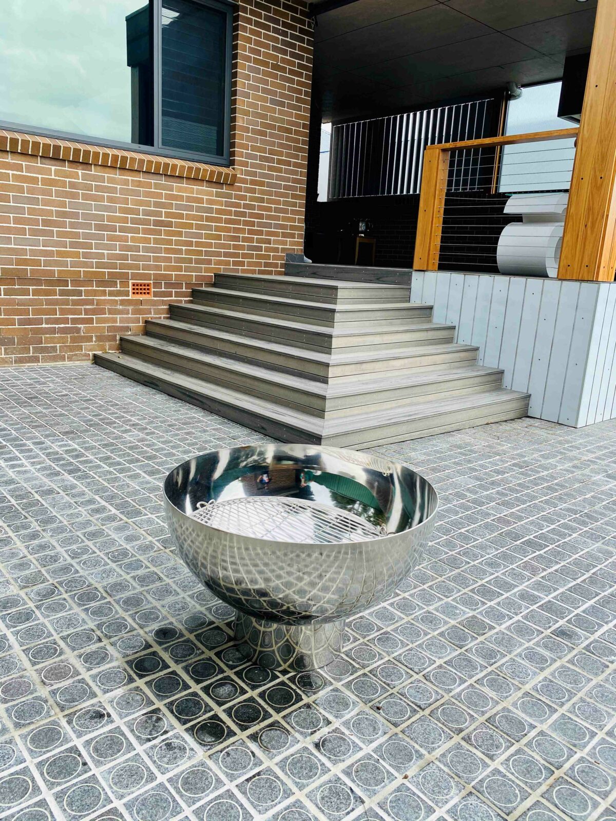 A stainless steel Goblet fire pit with a cooking grill inside, on a grey tiled courtyard below brick steps