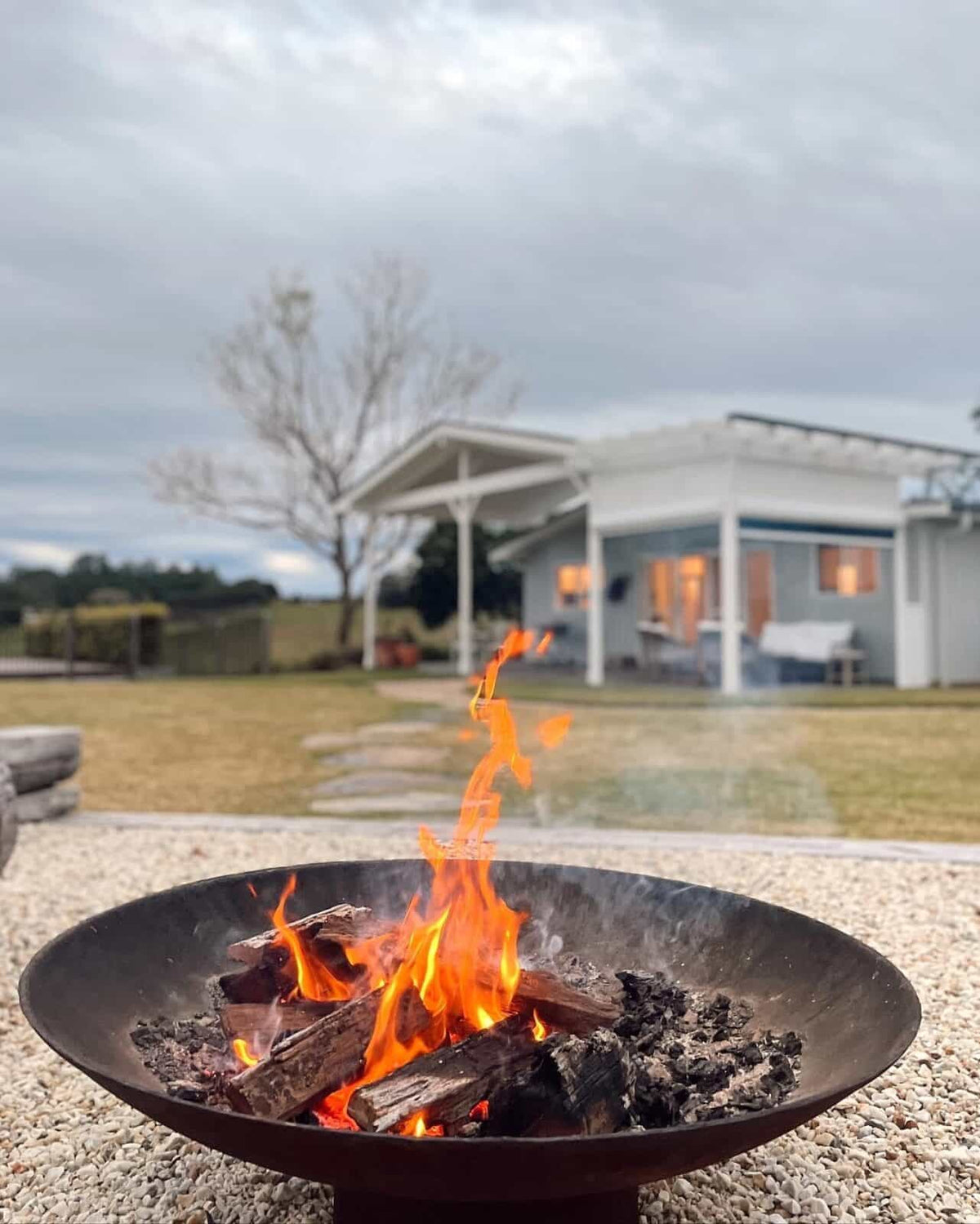 The Cauldron 1000 mm with a log fire burning on a gravel pad, with a white weatherboard house behind