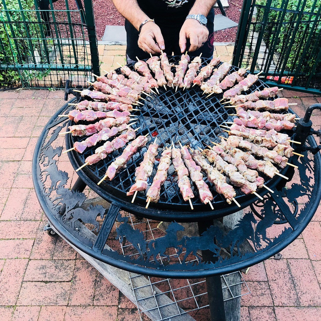 Meat skewers laid in a ring around the grill of a BBQ / Fire Pit with galloping horses cut into the rim, on a brick patio
