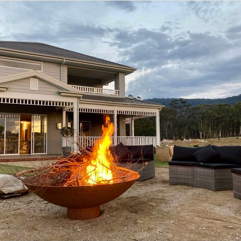 The Cauldron 1500 mm with a tall fire burning in front of a two-storey house with a verandah and outdoor lounges