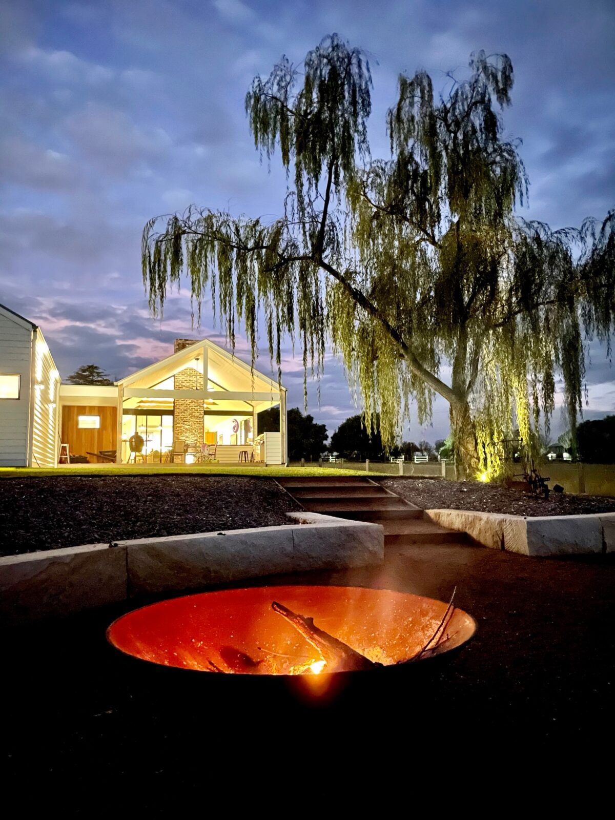 A Cauldron glowing with coals at night, with a lit house and a willow tree behind