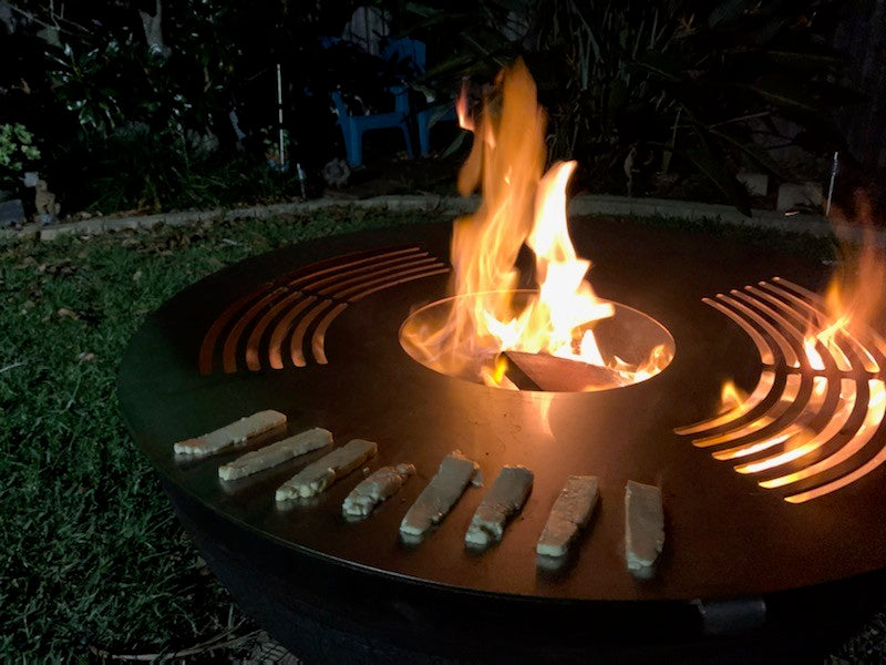 Food cooking on a stainless hot plate ring while a fire burns through the centre hole at night