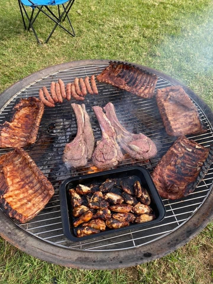 Lamb cutlets, ribs, sausages and a tray of chicken wings on a round stainless steel grill over a fire pit on the lawn