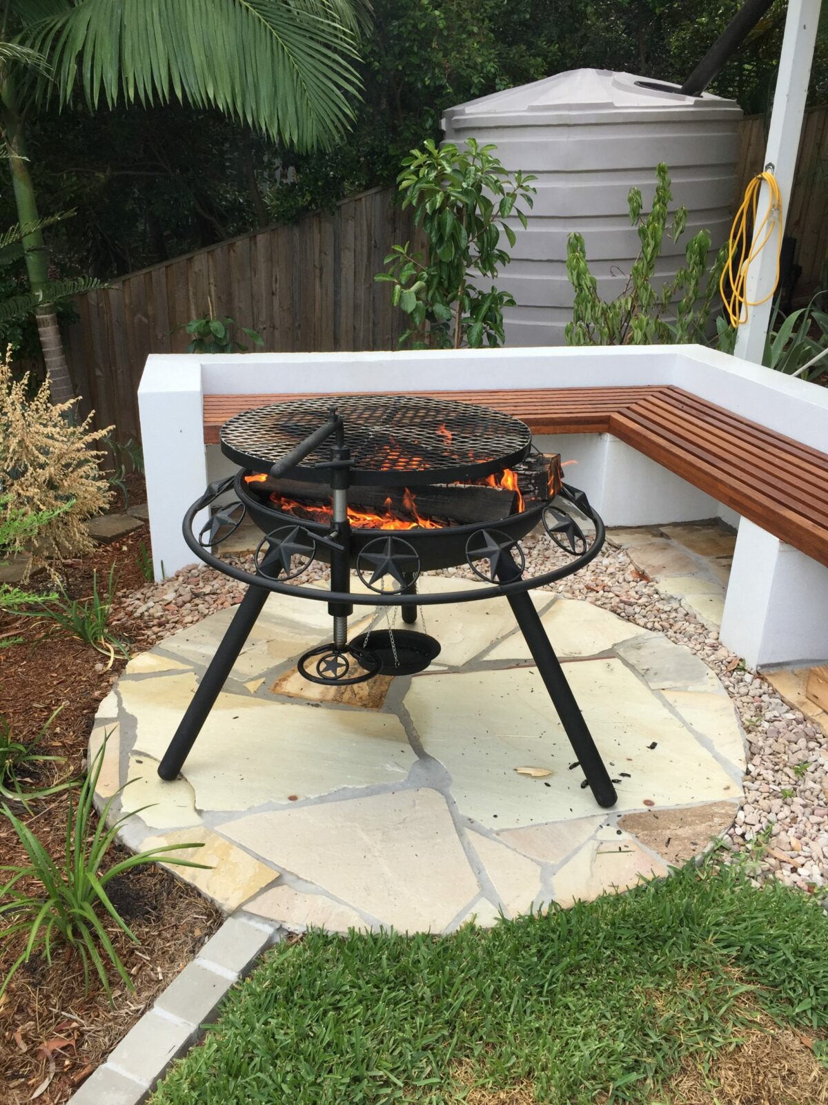 A BBQ / Fire Pit with stars cut into its rim, a fire burning under the grill, on a sandstone pad beside a built-in timber bench