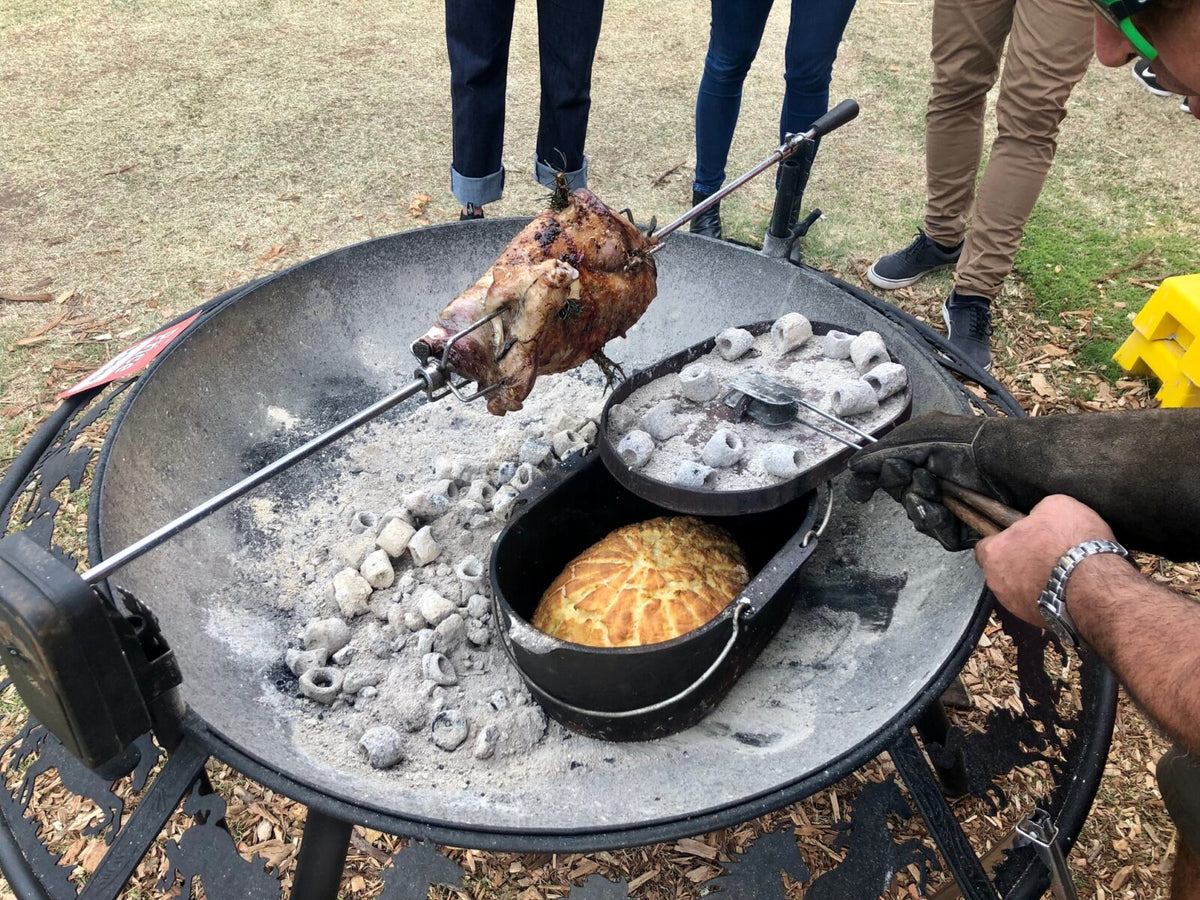 A lamb turning on a spit over white-hot heat beads in a BBQ / Fire Pit, a damper baking in a camp oven in the coals