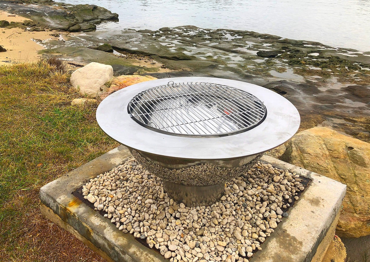 A stainless steel Teppanyaki fire pit with a grill in the centre of its wide top plate, on a bed of white pebbles beside a river