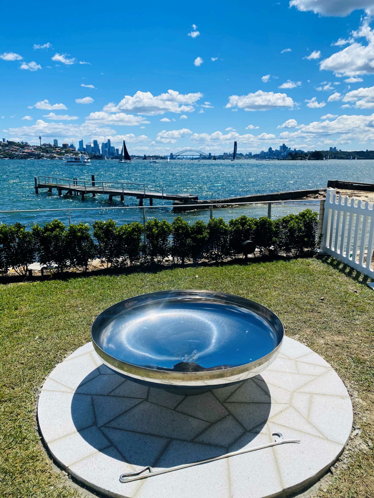 A mirror-polished stainless steel Cauldron on a round paved pad on a waterfront lawn, with the harbour and the city skyline behind