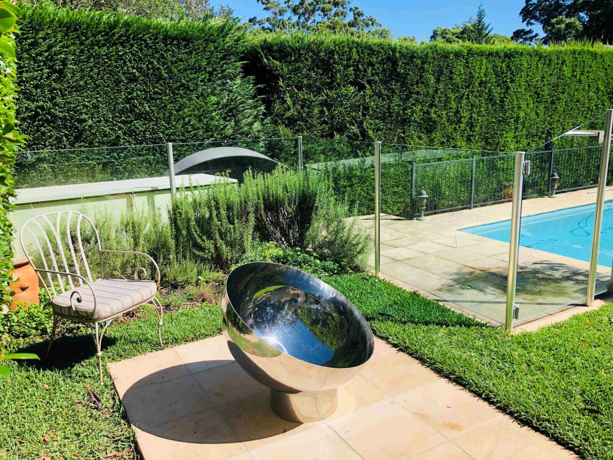 A mirror-polished stainless steel Goblet fire pit on a paved pad in a garden, with a hedge and a swimming pool behind