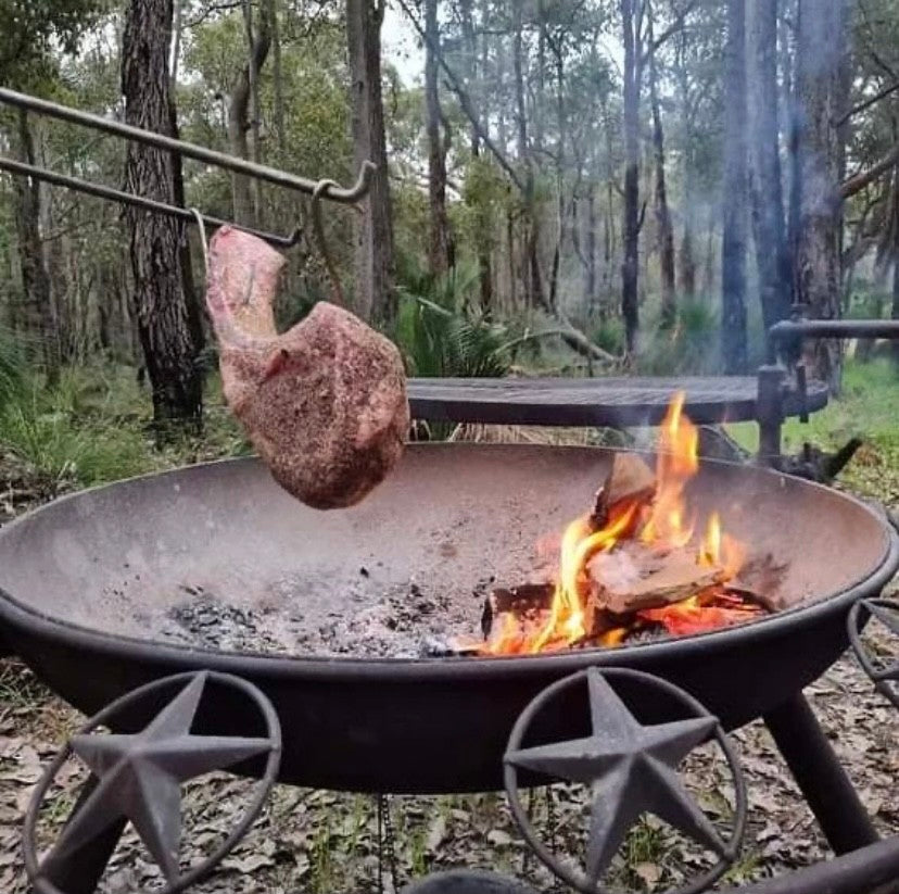 A tomahawk steak hanging over a wood fire in a BBQ / Fire Pit with stars on its rim, in the bush