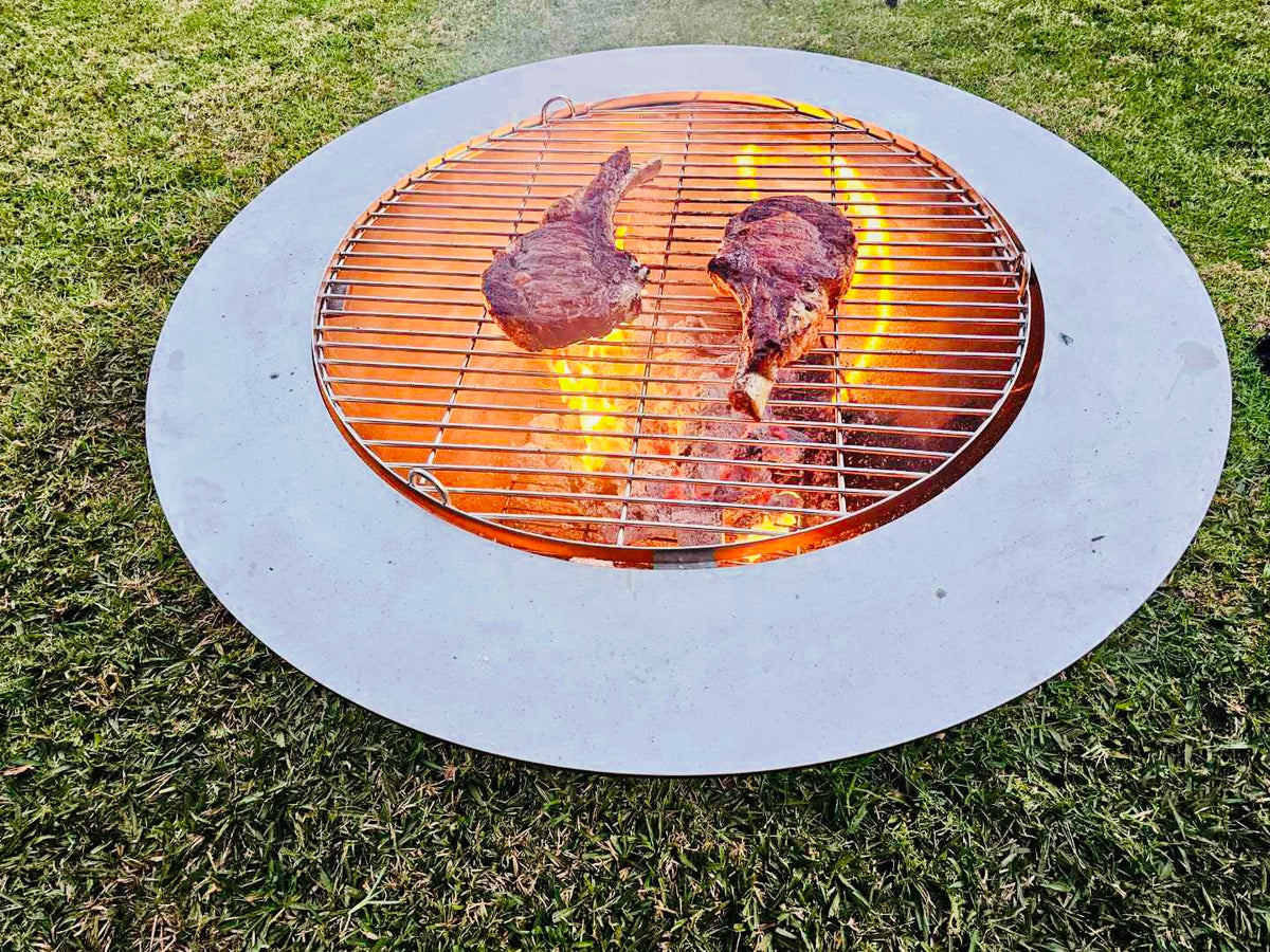 Two steaks on the round grill in the centre of a Teppanyaki fire pit, flames under the grill and a wide flat top plate around it, on a lawn