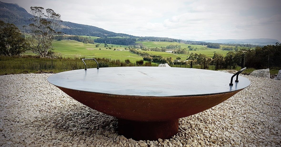 A steel fire pit lid with two handles sitting on a cast iron Cauldron, green hills behind