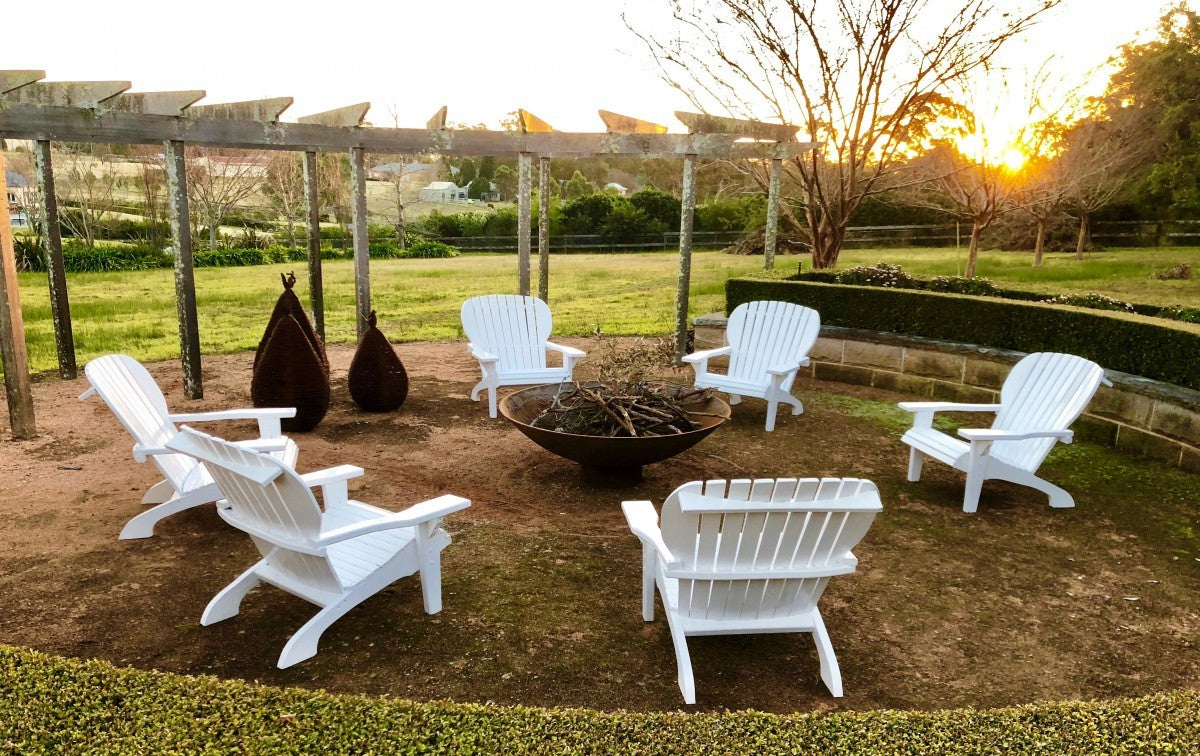 White Adirondack chairs around a cast iron Cauldron on a lawn at sunset, with a timber pergola behind
