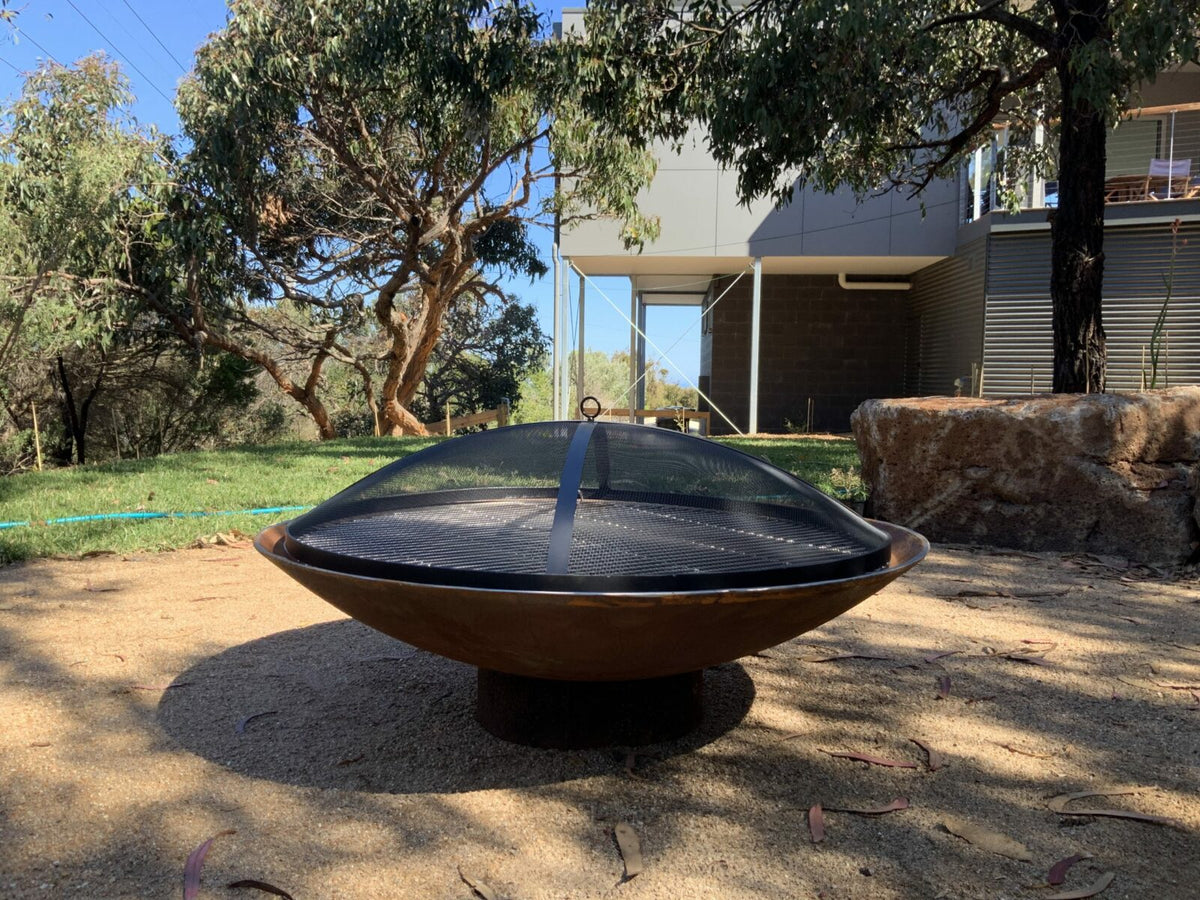 A domed mesh ember screen sitting on a cast iron Cauldron in a backyard