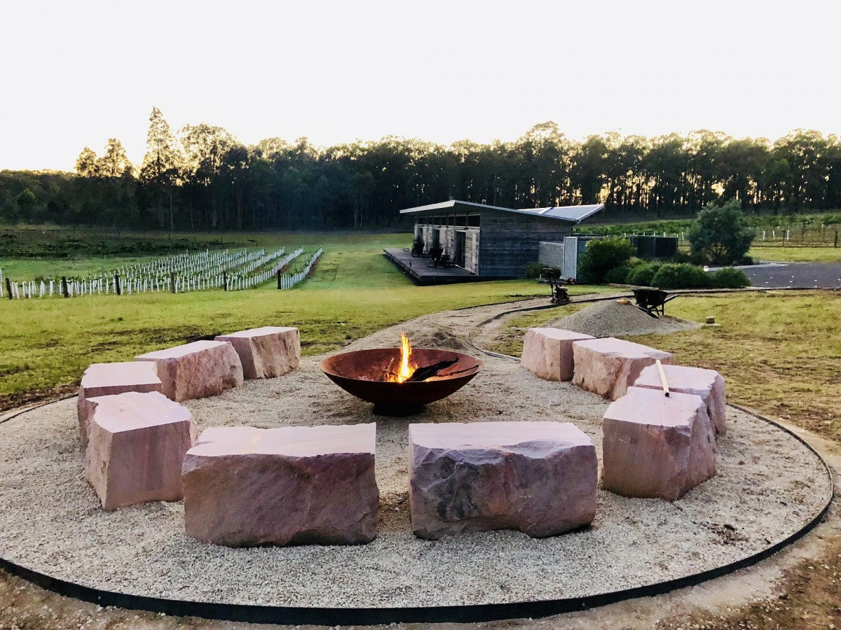 A Cauldron with a small fire going, ringed by sandstone block seats, with a vineyard and a timber shed behind