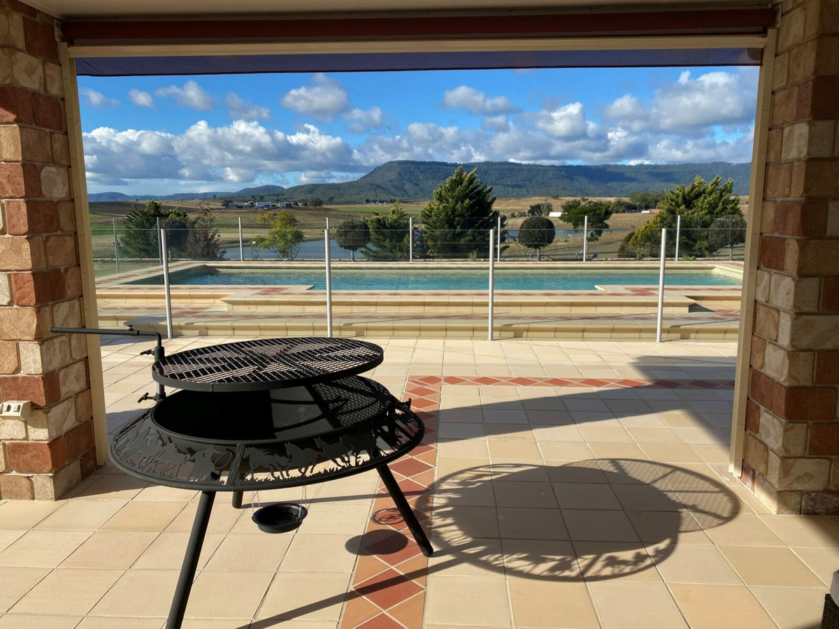 A BBQ / Fire Pit on a tiled patio in the morning sun, its grill still on, with a pool and hills behind