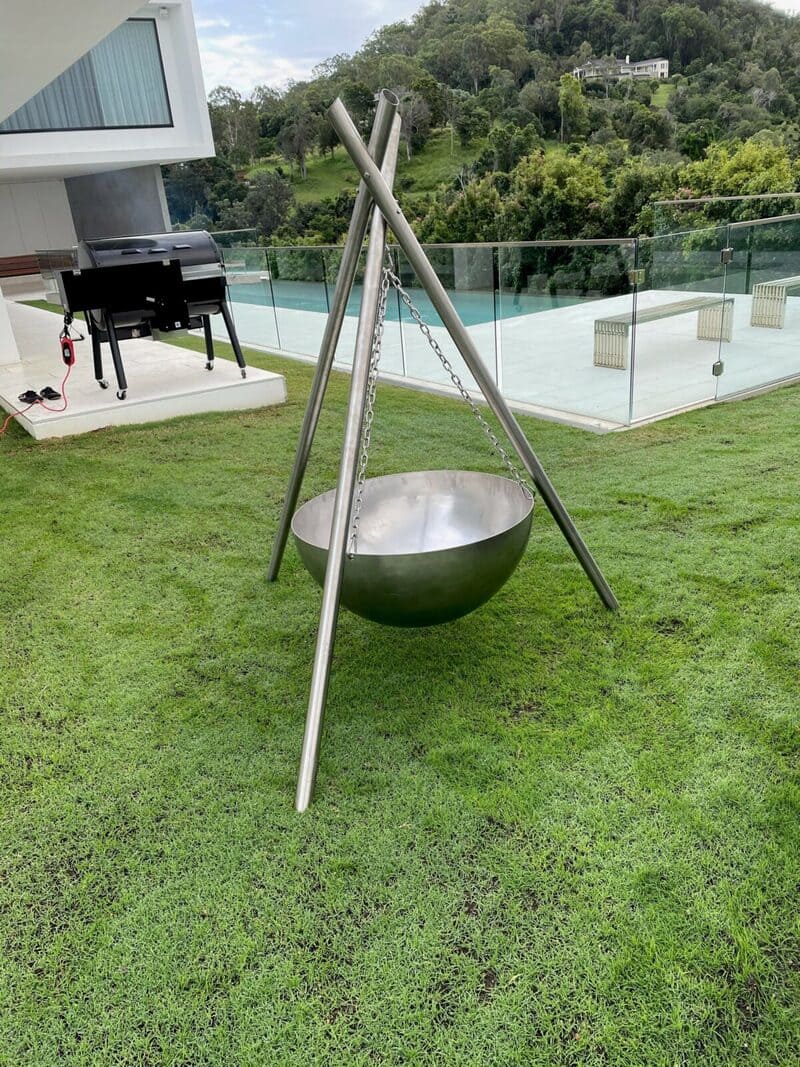 A stainless steel Tripod fire pit on a lawn, its bowl hanging above the grass, with a glass pool fence behind