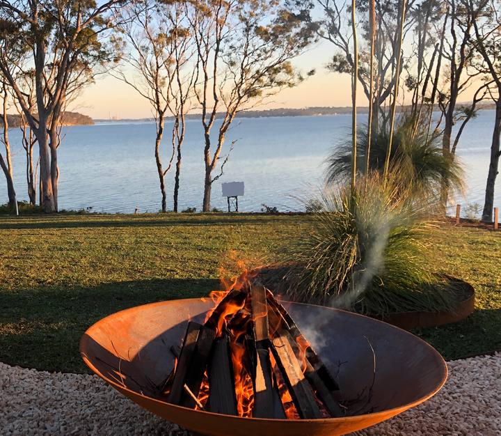 A log fire burning in a cast iron Cauldron on a gravel path at golden hour, with a lake and gum trees behind