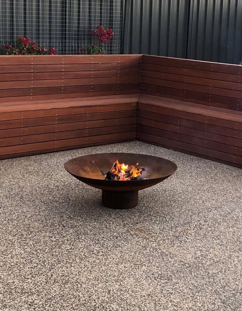 A cast iron Cauldron with a small fire going in a pebble courtyard, beside an L-shaped timber bench and a black sun lounger